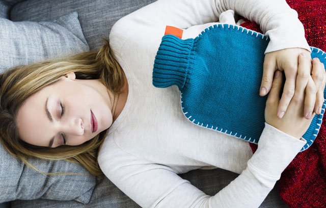 Frau liegt auf dem Sofa mit einer Wärmeflasche auf dem Bauch 